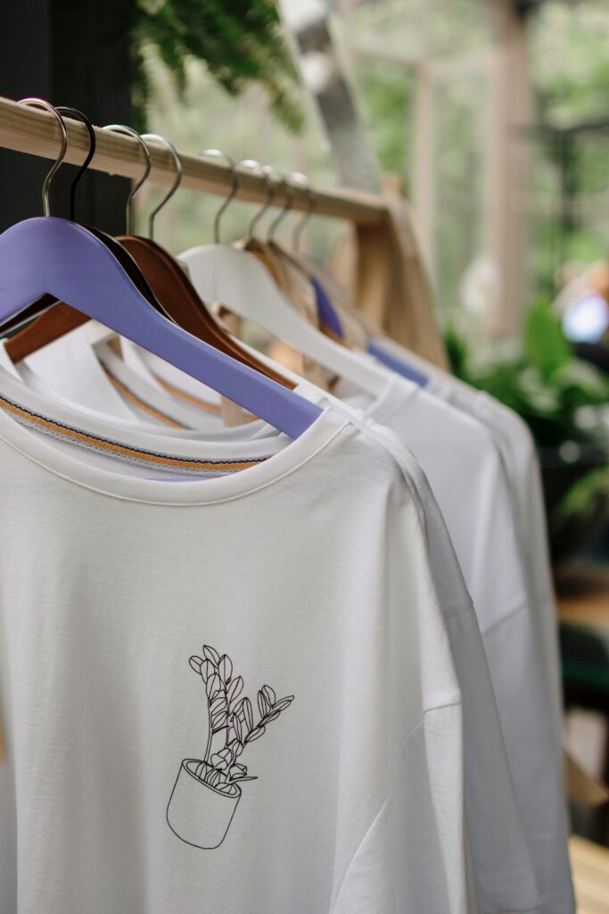 White t-shirts with simple plant design hang on wooden rack indoors, showcasing minimalist fashion.